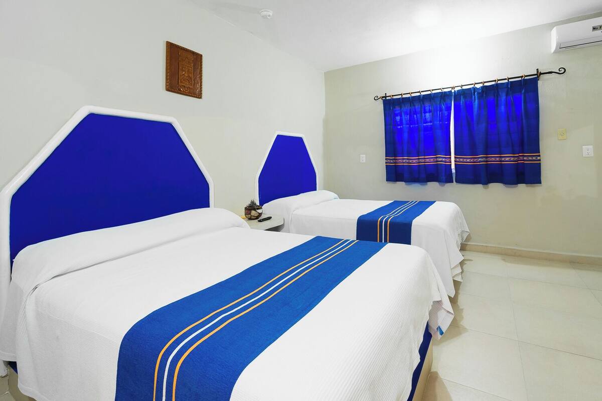Habitación de hotel con dos camas, cada una con cabecero azul y ropa de cama blanca. Hay una ventana con cortinas azules, una mesita con lámpara y aire acondicionado de pared.