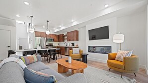 A modern living room with a gray sofa, a wooden coffee table, and a mustard-colored armchair. A kitchen with wooden cabinets and a dining area with a white table and chairs is visible in the background.