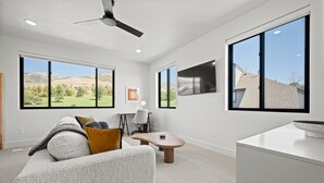 A modern living room with a large window, a ceiling fan, a flat-screen TV, and a cozy seating area.