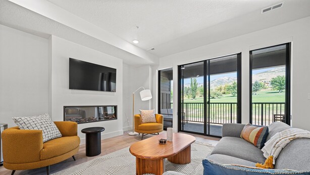 A modern living room with a sofa, armchair, and coffee table, a flat-screen TV mounted on the wall, and a view of a green landscape through large sliding doors.