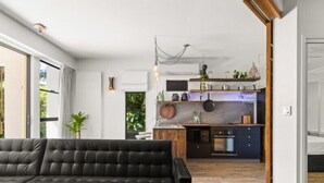 A modern living room with a black leather sofa, a wooden coffee table, and a large window providing natural light. A kitchen with a central island and built-in appliances is visible in the background. - Casuarina Apartment 200m from the Beach (Casuarina)