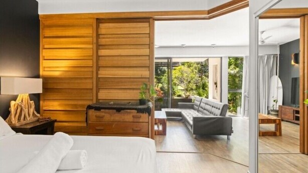 A modern hotel room with a large bed, wooden paneling, a sofa, and a view of greenery through large windows. - Casuarina Apartment 200m from the Beach (Casuarina)