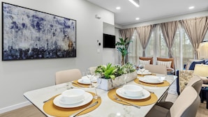 A modern dining area with a marble table, white plates, and gold placemats. There is a large abstract painting on the wall and a plant in a planter.