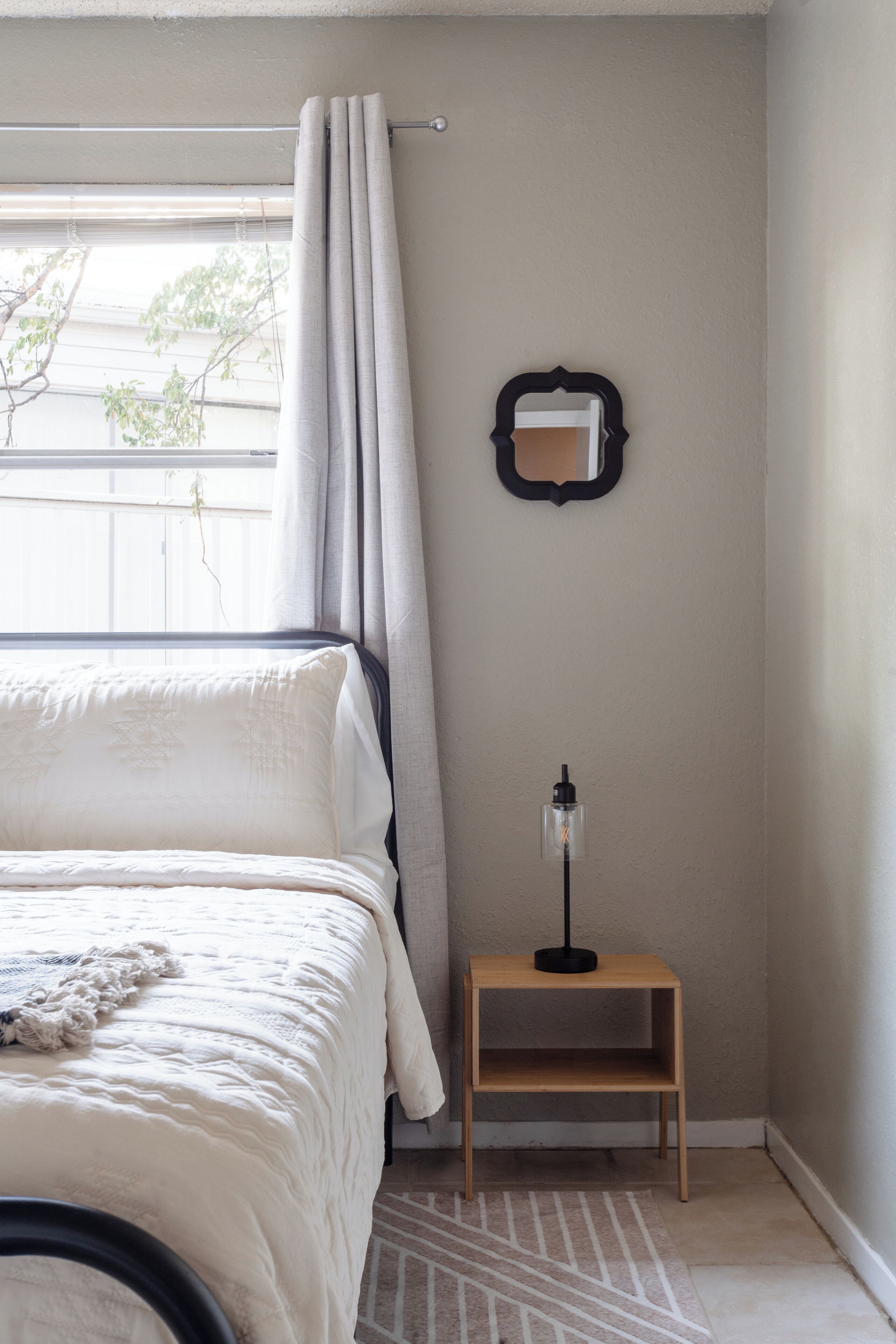 A bedroom with a bed, a nightstand, a wall-mounted mirror, and a window with curtains.