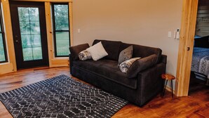 Ein gemütliches Zimmer mit einem dunkelbraunen Sofa, einem gemusterten Teppich und einer Holztür, die auf einen Balkon mit Blick auf Grünflächen führt.
