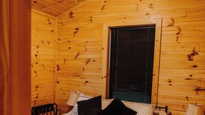 Ein Schlafzimmer in einer Holzhütte mit einem Bett, zwei Nachttischlampen, einem Deckenventilator und einem Fenster mit Jalousien.