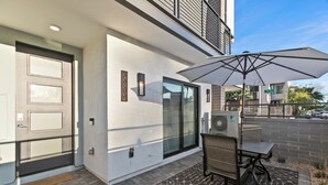 Une entrée moderne avec une porte en verre, un patio couvert doté d’une table et de chaises, et un grand parasol.