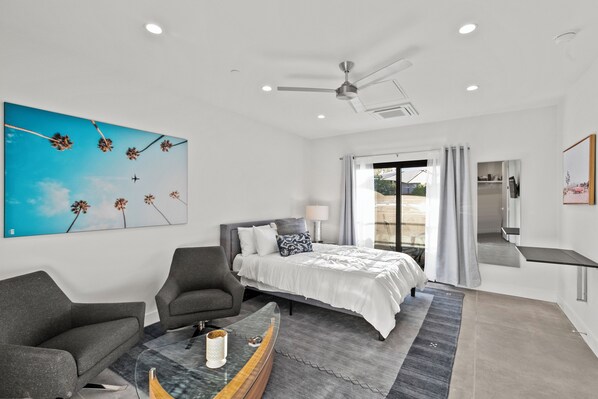 A modern bedroom with a bed, two armchairs, a coffee table, a ceiling fan, and a large window with curtains.