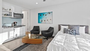 Une chambre d’hôtel moderne dotée d’une kitchenette, d’une table basse en verre, de deux fauteuils, d’un lit avec une literie blanche et d’un ventilateur de plafond.