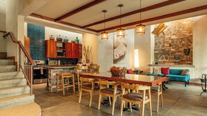 Une cuisine spacieuse avec des meubles en bois, un coin repas avec une longue table et des chaises, une cheminée en pierre et un escalier menant à l’étage supérieur.