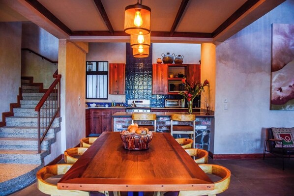 A spacious dining area with a long wooden table, surrounded by wicker chairs, and a central light fixture.