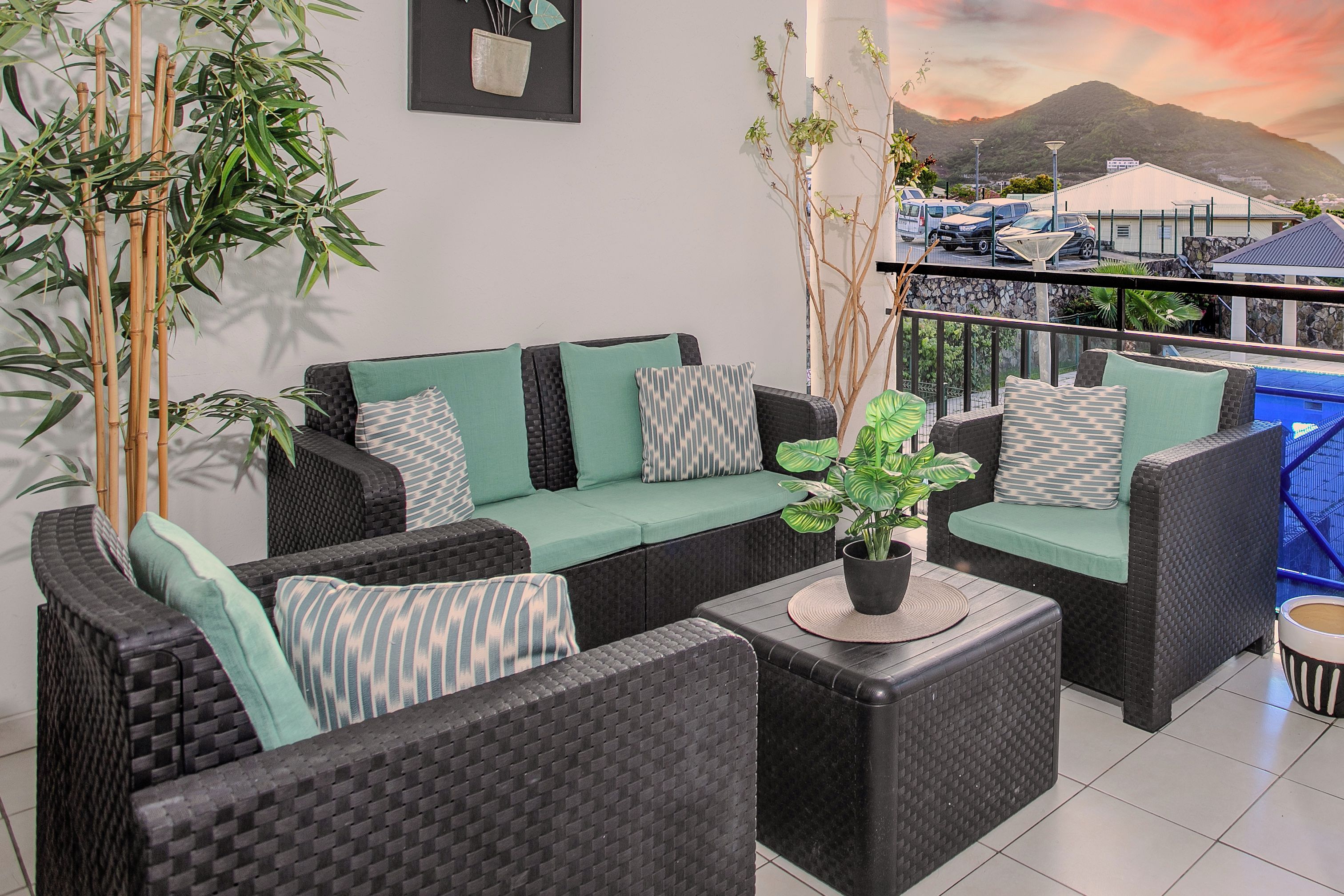A balcony with wicker furniture, a potted plant, and a view of a mountain and parked cars.