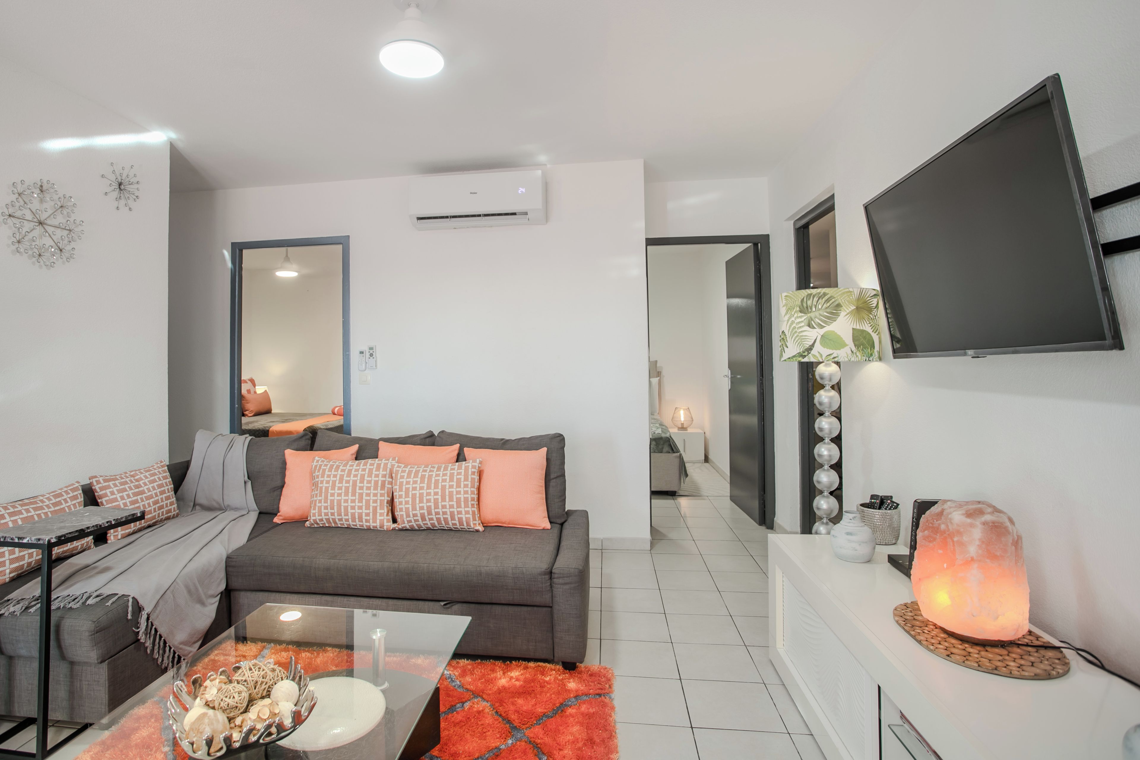 Ein modernes Wohnzimmer mit einem grauen Sofa, orangefarbenen Kissen, einem Glastisch und einem an der Wand befestigten Fernseher.