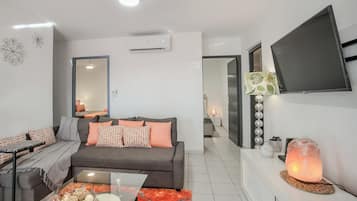 Un salon moderne avec un canapé gris, des coussins orange, une table basse en verre et un téléviseur fixé au mur.