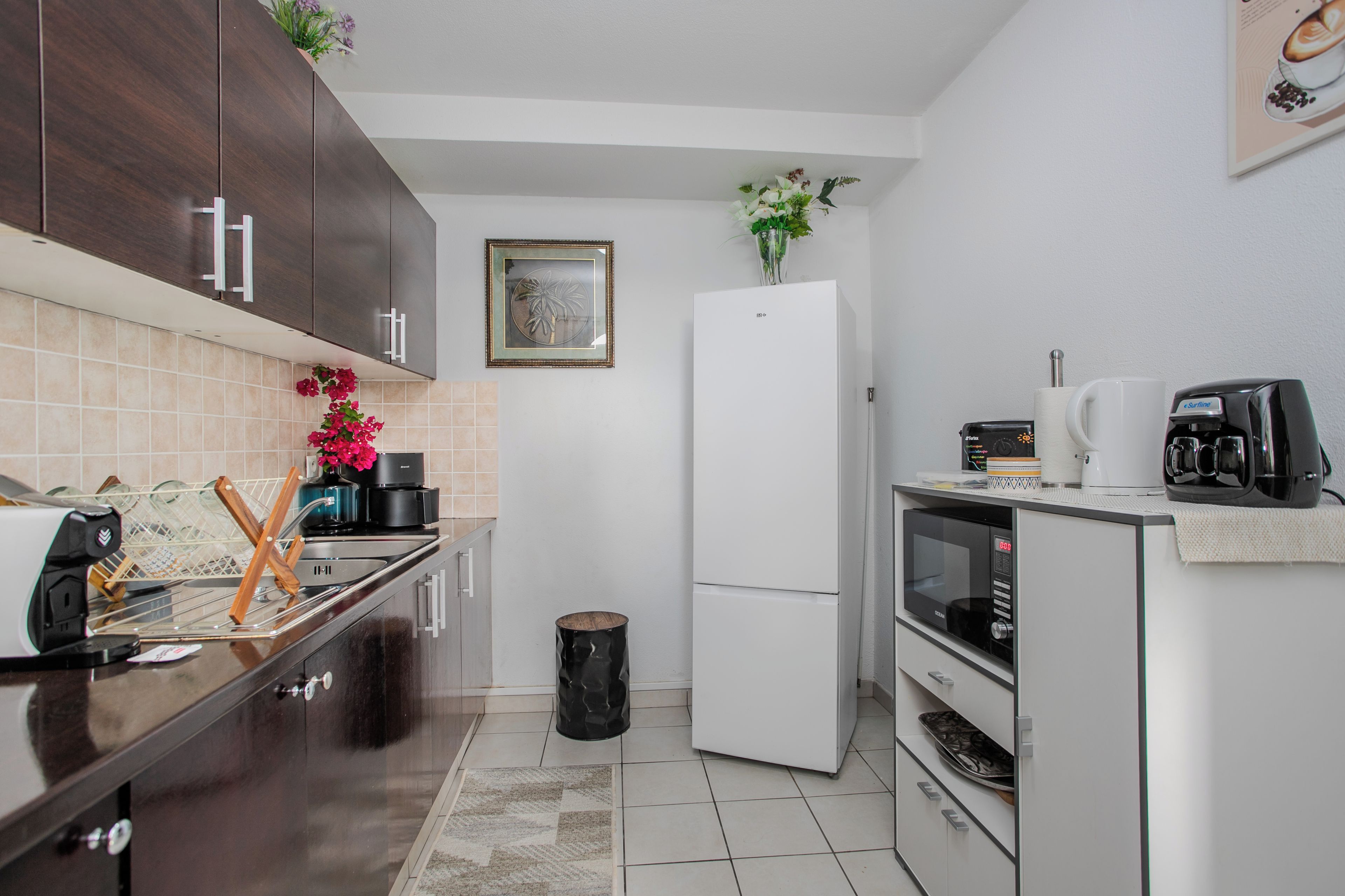 Cocina compacta con armarios de madera oscura, un refrigerador blanco, una cafetera y un microondas.