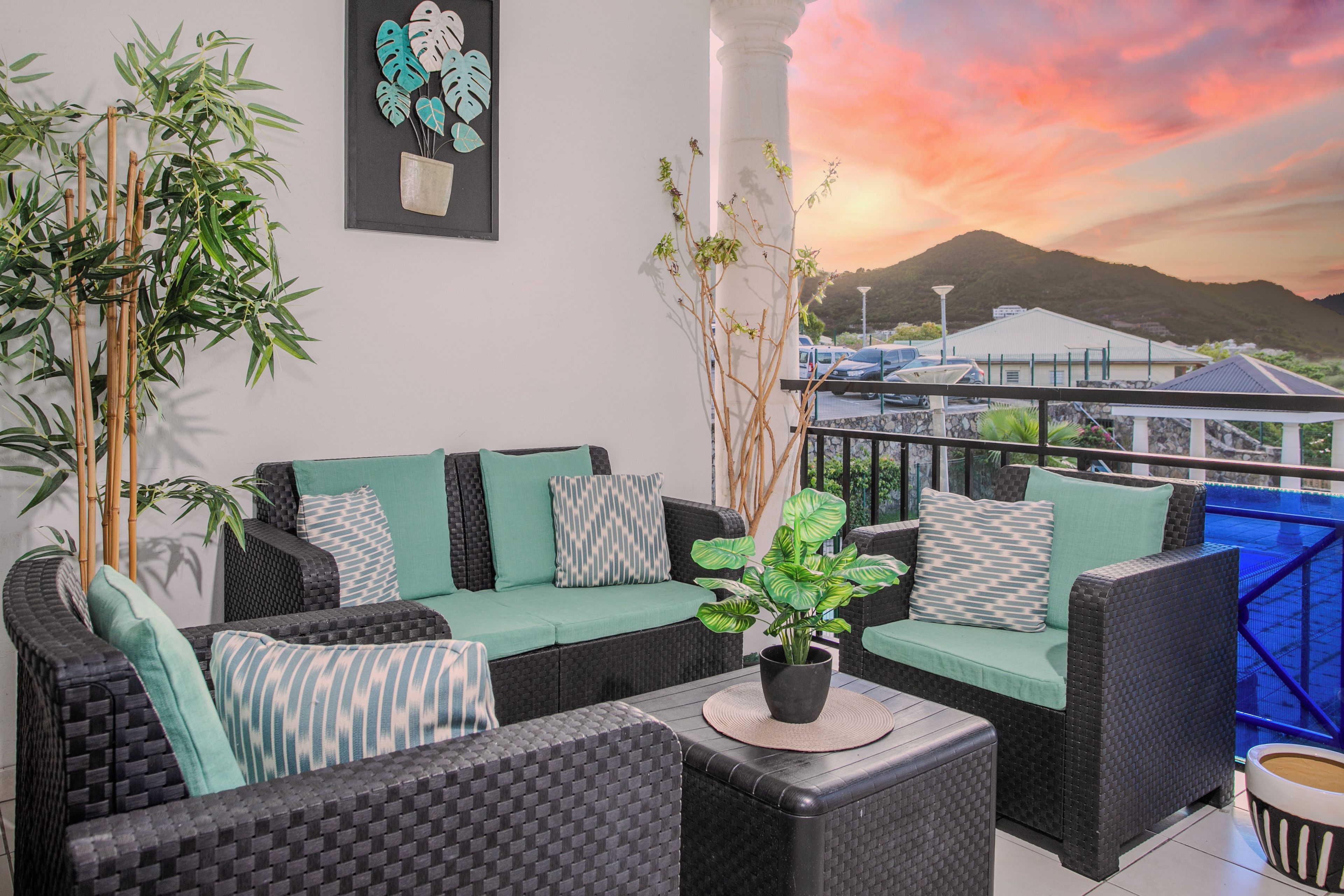 A balcony with wicker furniture, a potted plant, and a view of a mountain at sunset.