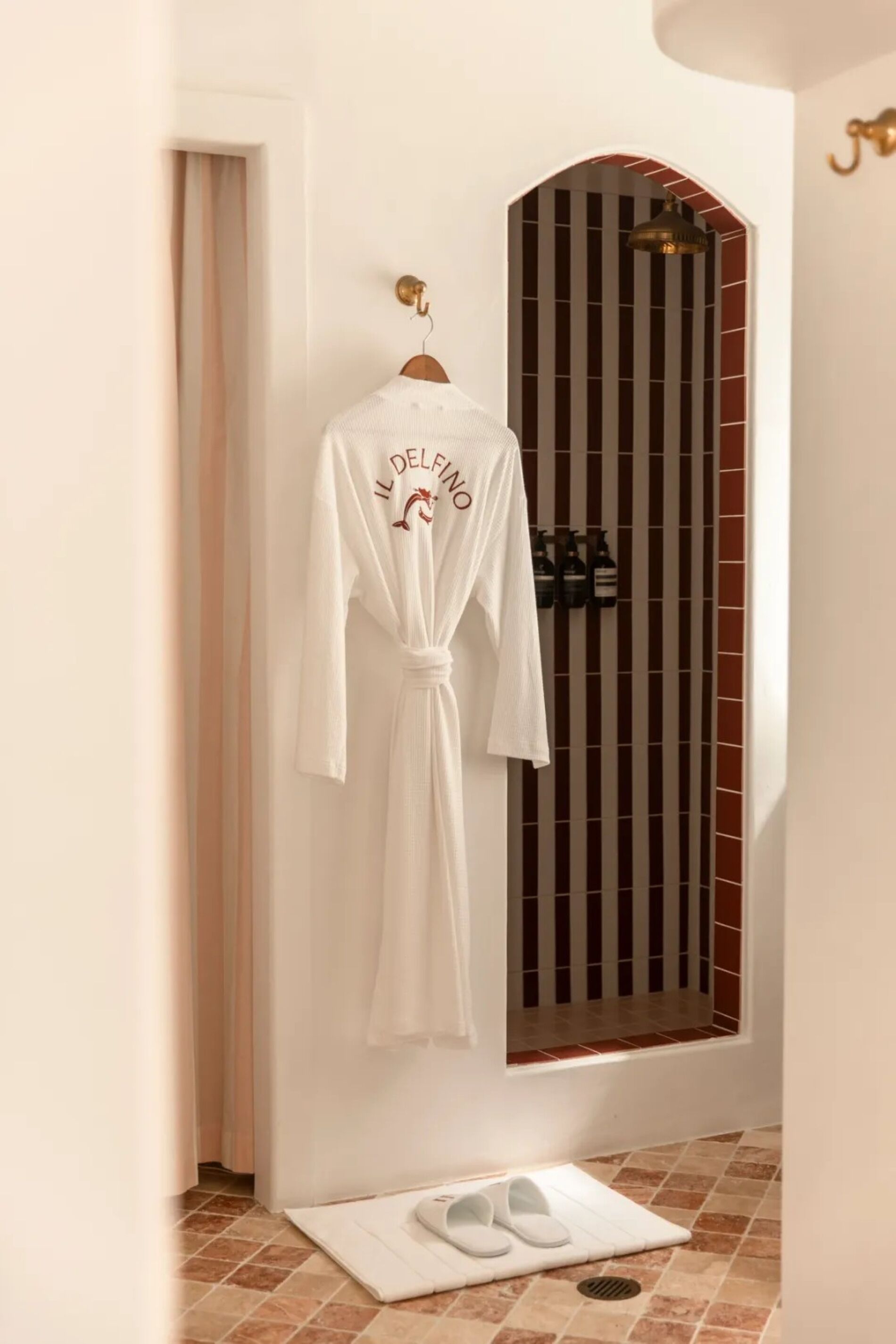 A white robe hanging on a wooden hanger in a bathroom with a tiled floor and a shower area with a striped glass door.