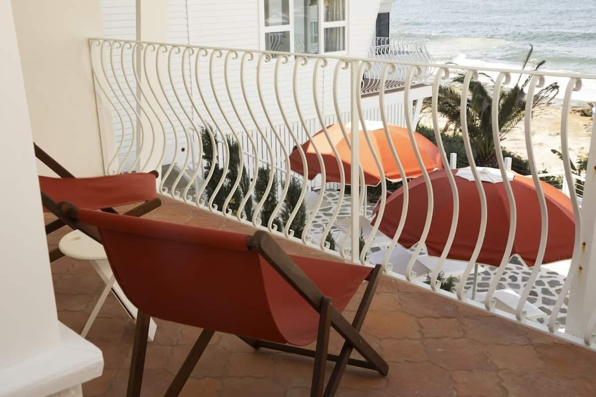 En balkon med gelænder i hvid metal, en rød pude stol, samt udsigt over stranden og havet.