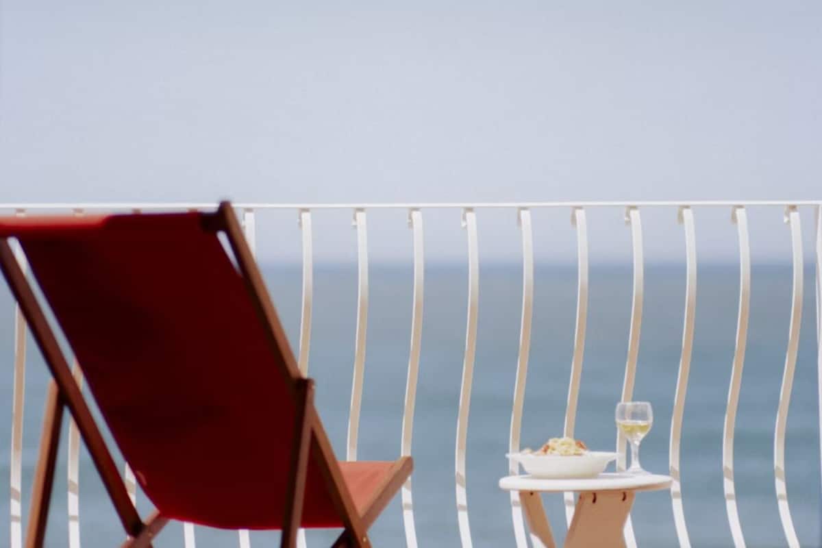 En rød stol og et lille bord med en tallerken og et glas hvidvin på en balkon med udsigt til havet.