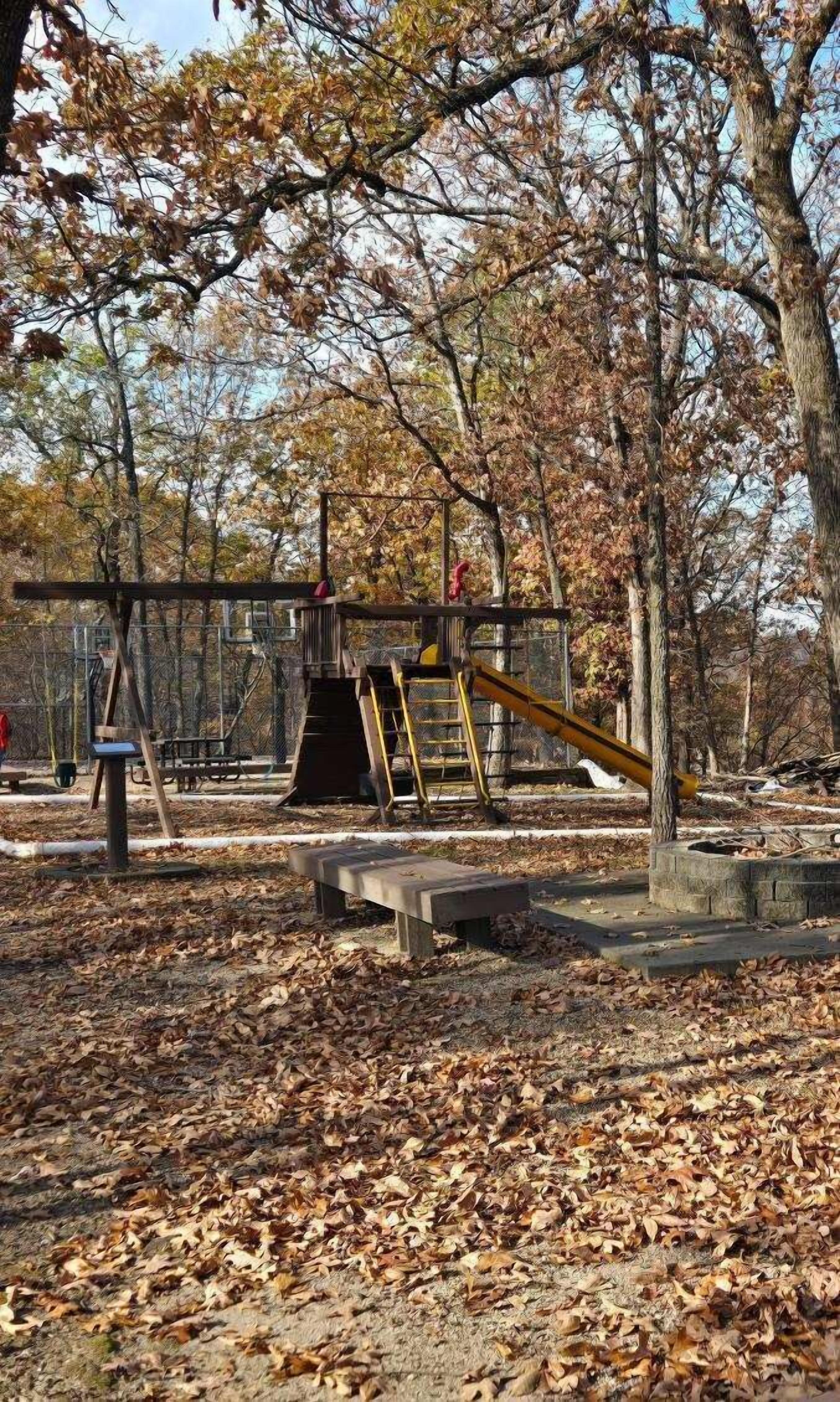 A playground with a swing set, slide, and climbing structure surrounded by trees and fallen leaves.