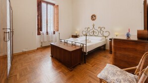 A bedroom with a wooden floor, a bed with a metal frame, a wooden chest of drawers, a chair, and a window with curtains. - La Margherita by MMega (Coverciano)
