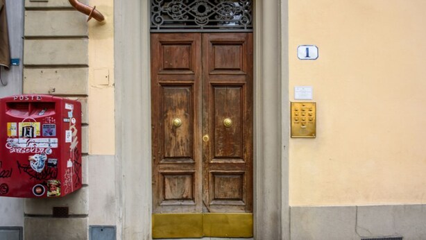 A double wooden door with brass handles, a red mailbox, and a yellow intercom on a beige wall.