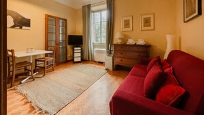 A room with a wooden floor, a red sofa, a television, a table with chairs, a window with curtains, and a wall with framed pictures.