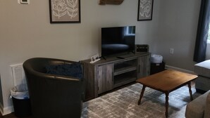 A living room with a large flat-screen TV, a dark-colored sofa, a wooden coffee table, and a patterned rug. - Curve Cottage located on The Rattlesnake 192 just outside of Somerset.  (Somerset)