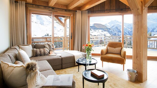 Una sala de estar acogedora con sofá, sillón y mesa de centro, ventanas amplias con vistas a la montaña y vigas de madera en el techo.