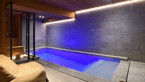 Una piscina interior moderna con borde iluminado en azul, techo de madera y paredes de piedra.