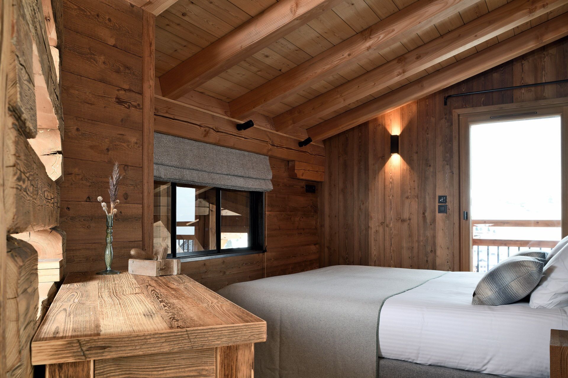 A wooden cabin room with a bed, a small table, a window, and a wall-mounted light fixture.
