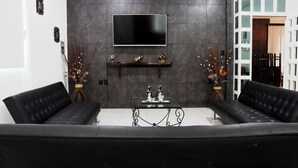 A modern living room with a black leather sofa, a flat-screen TV mounted on a dark tiled wall, and decorative items on a shelf below. - Casa Santa Cecilia (Tehuacán)