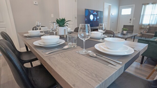 Una mesa de comedor dispuesta con platos, cuencos y cubiertos blancos, y un televisor al fondo.