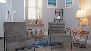 Sala de estar con dos sillones grises, una mesita redonda con una lámpara y una mesa baja con copas y botellas de vino. Hay cuadros enmarcados en la pared y una ventana con persianas.