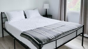 A neatly made bed with a white duvet and a patterned throw, a round wall decoration, a bedside table with a lamp, and a window with curtains.
