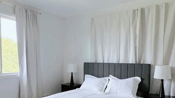 A neatly made bed with white linens in a room with a large window, a dark headboard, and two bedside tables with lamps.