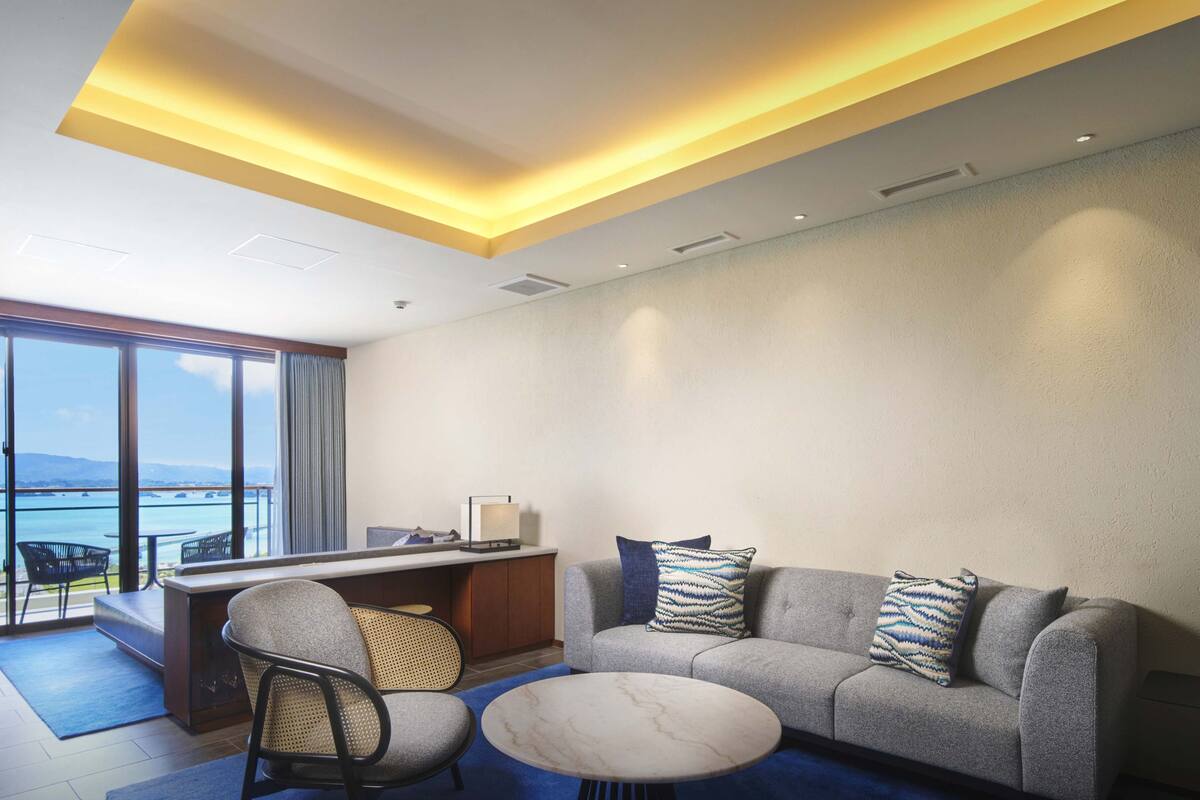 Une chambre d’hôtel moderne dotée d’une grande fenêtre donnant sur la mer, d’un canapé avec des coussins à motifs, d’une petite table ronde et d’un bureau avec une lampe.