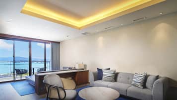 A modern hotel room with a large window offering a view of the sea, a sofa with patterned cushions, a small round table, and a desk with a lamp.