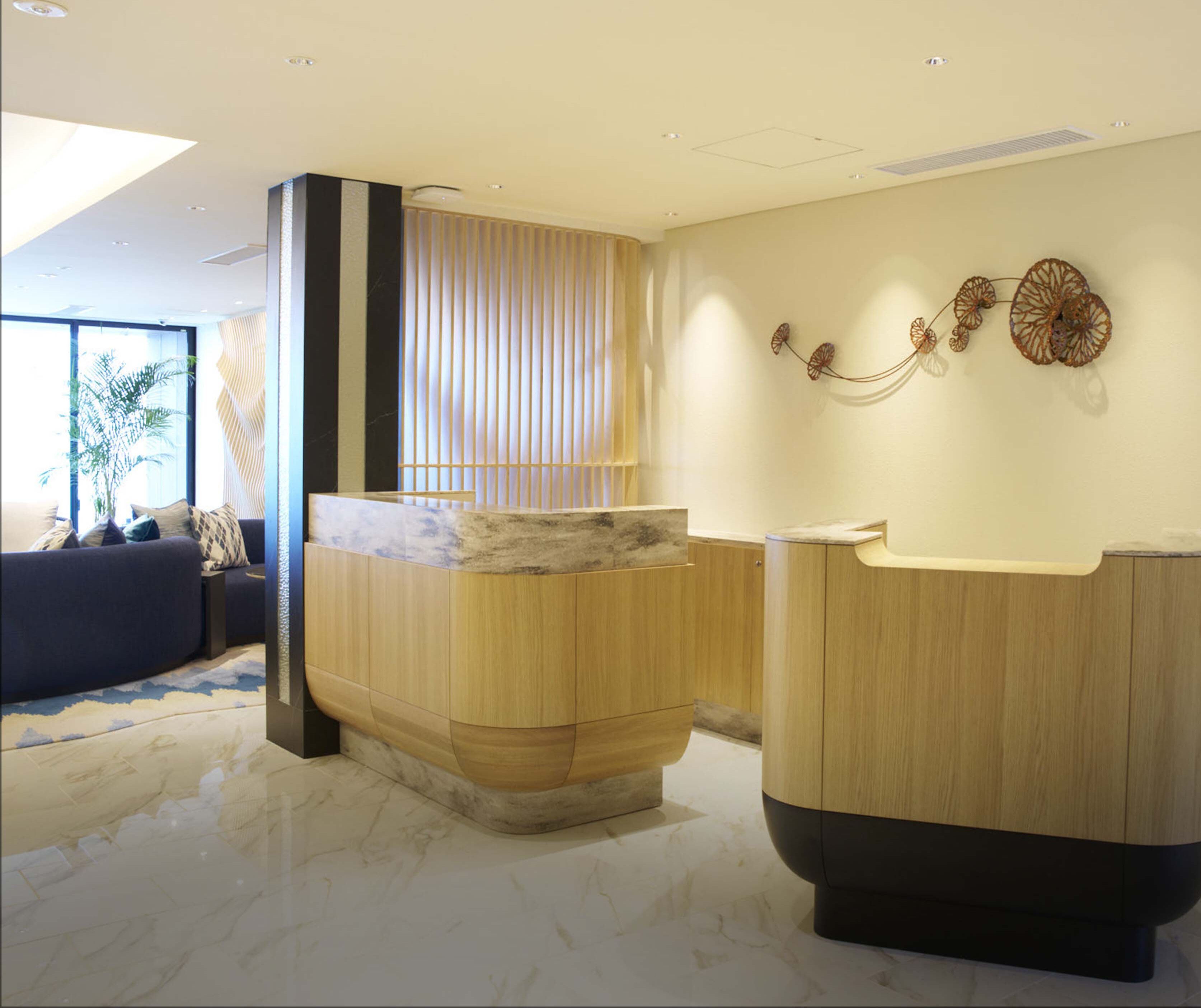 A modern reception area with a wooden reception desk, a seating area with a sofa and a potted plant, and a decorative wall piece.