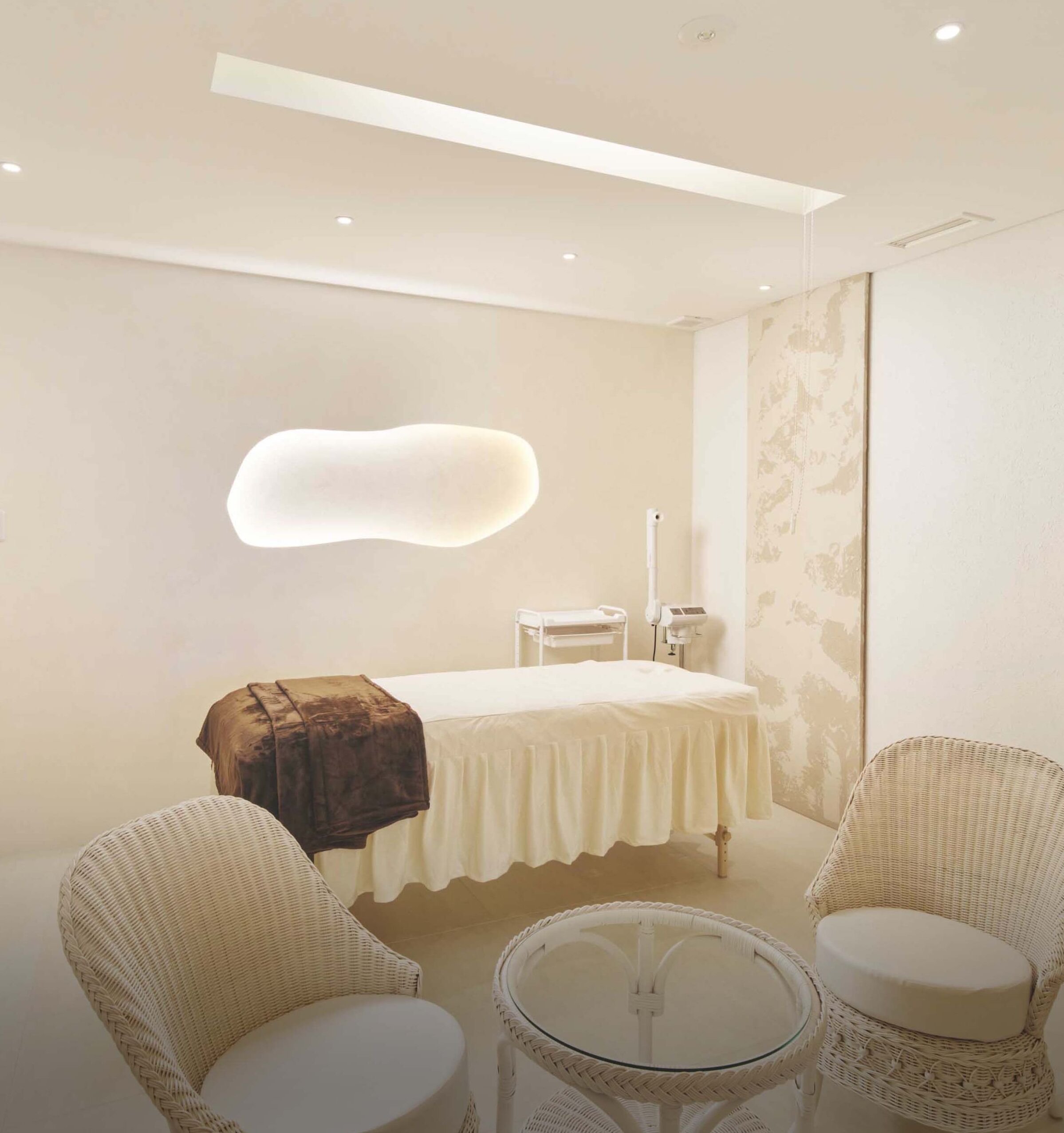 A room with a bed, two wicker chairs, a small round table, and a wall-mounted light fixture.