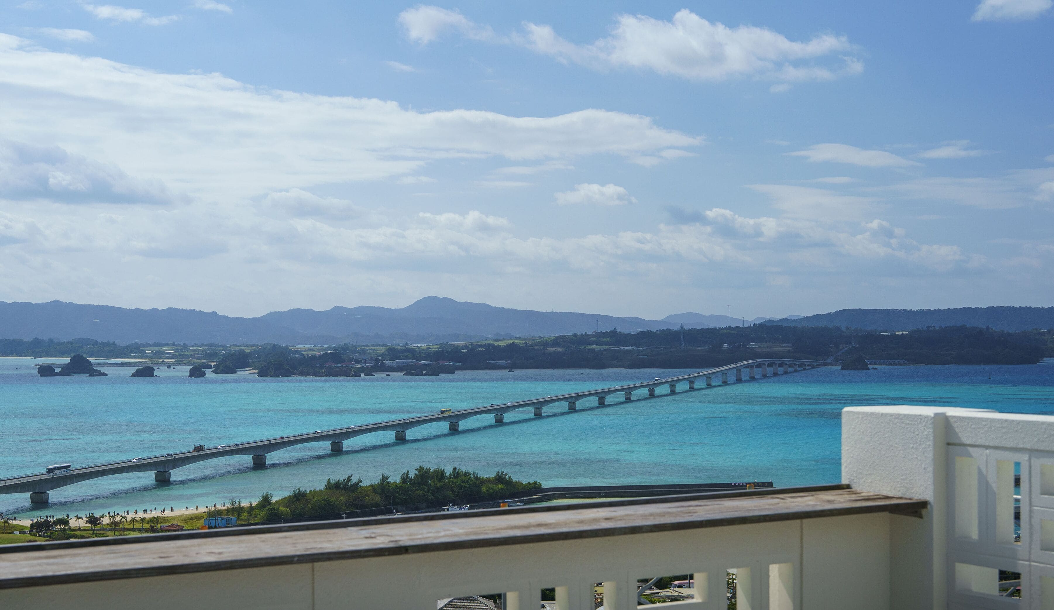 A coastal view with a long bridge over turquoise waters, a beach, and distant mountains.