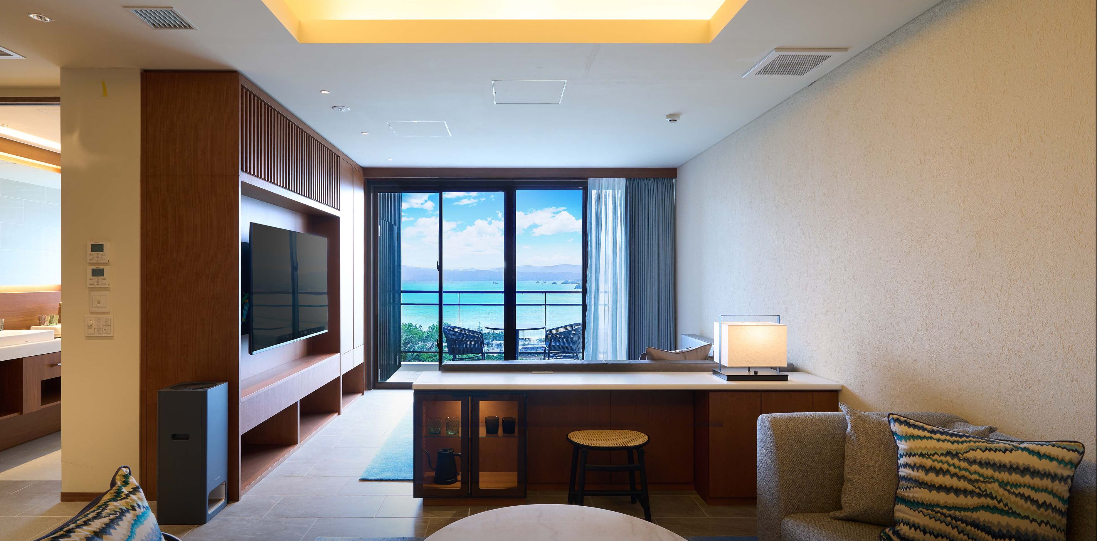 A modern hotel room with a large window offering a view of the sea, a flat-screen TV, a desk with a lamp, and a sofa with patterned cushions.