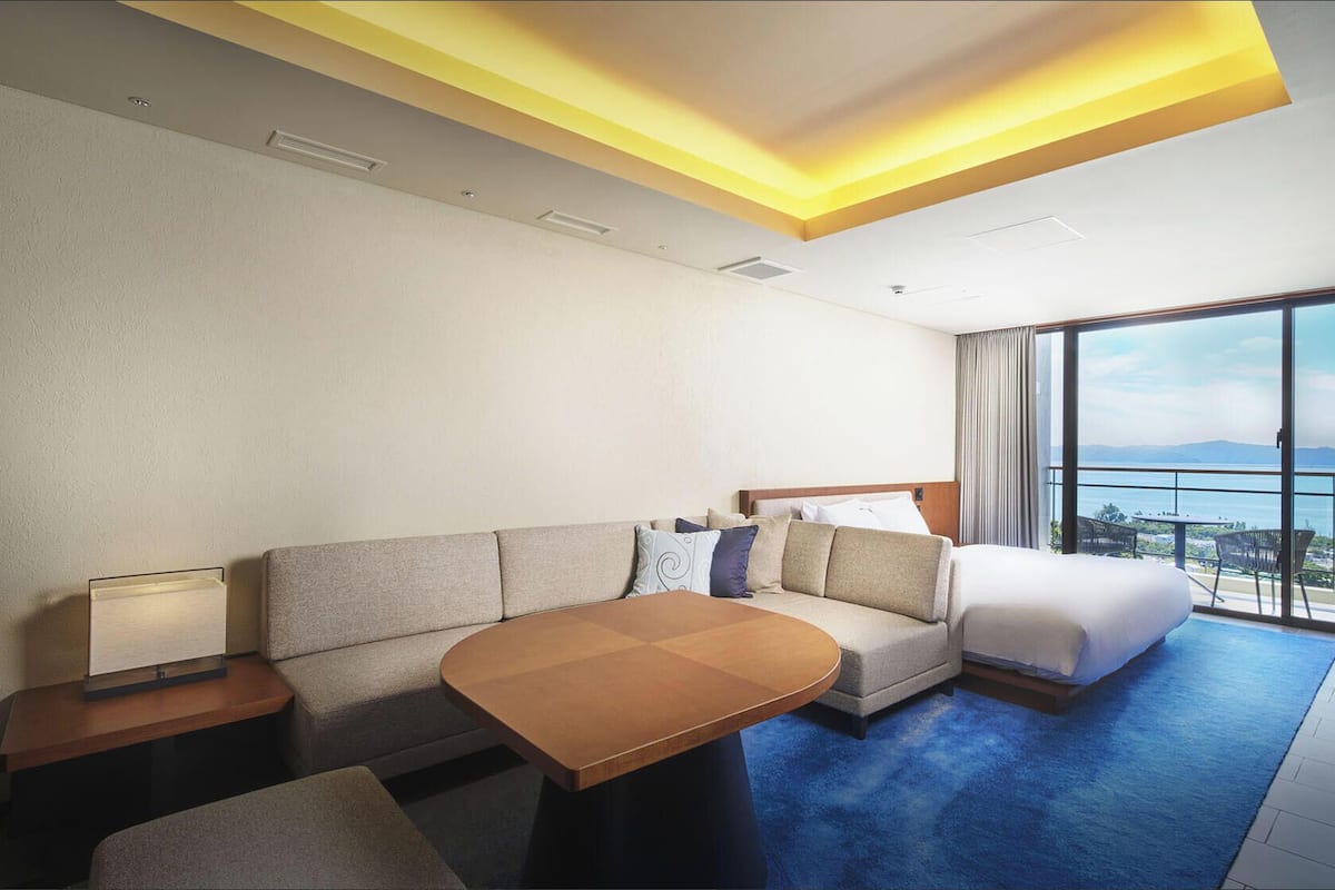A hotel room with a sofa, a bed, a table, and a view of the sea.