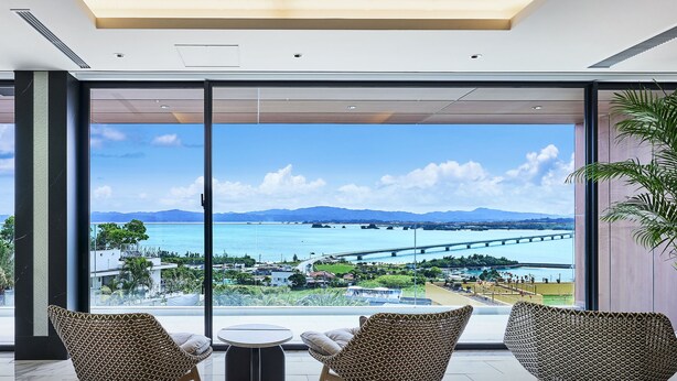 A modern hotel room with large windows offering a view of a coastal city, a bridge, and mountains. - BATON SUITE Okinawa Kouri Island (Nakijin)