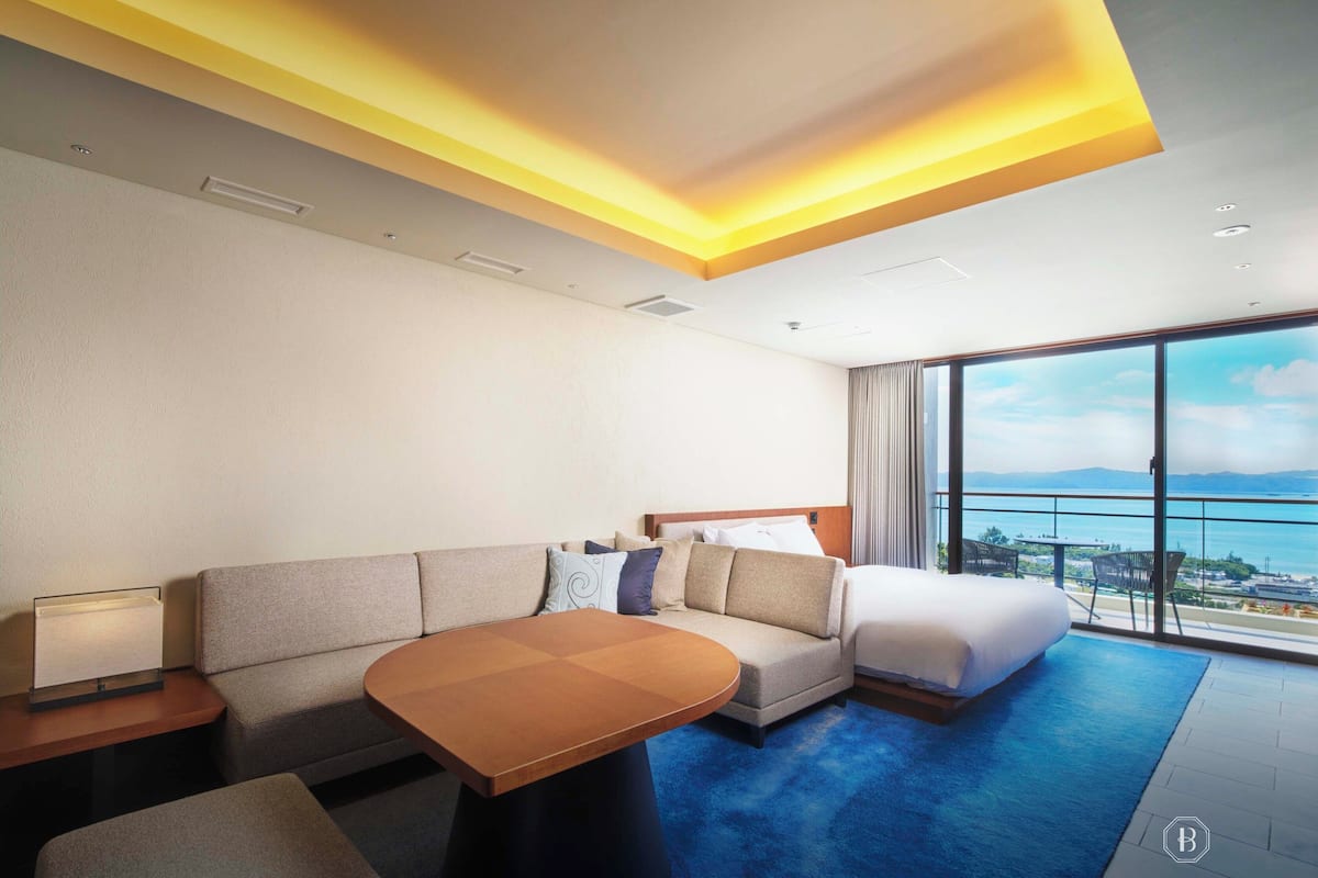 Une chambre d’hôtel moderne, équipée d’un canapé, d’un lit, d’une petite table et d’une grande fenêtre donnant sur la mer.