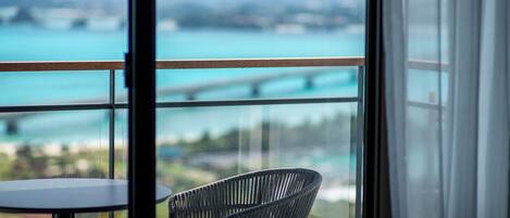 A balcony with a view of a bridge over turquoise water, a wicker chair, and a table.