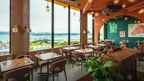 A restaurant with large windows offering a view of a bridge and water, wooden tables and chairs, hanging pendant lights, and a green accent wall with artwork. - BATON SUITE Okinawa Kouri Island (Nakijin)