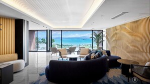A modern living room with a curved sofa, a round coffee table, and a seating area with chairs. A large window offers a view of the ocean and mountains. - BATON SUITE Okinawa Kouri Island (Nakijin)