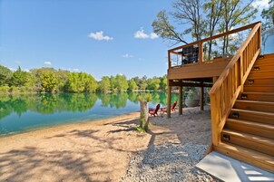 Una terraza de madera con una escalera que conduce a un área cubierta, con vistas a un lago tranquilo y árboles al fondo.