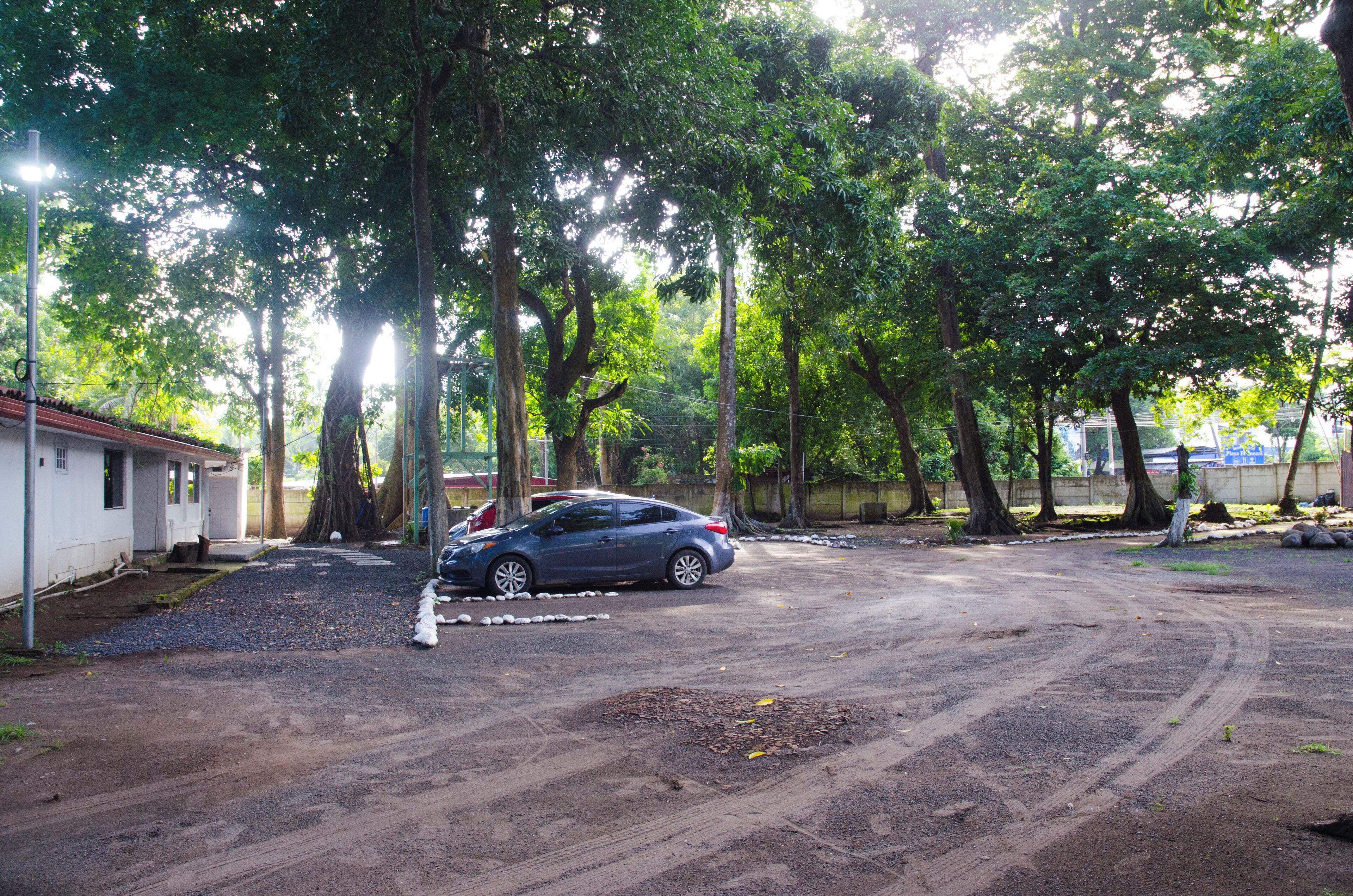 Un estacionamiento con suelo de tierra, rodeado de árboles y un edificio de techo rojo.